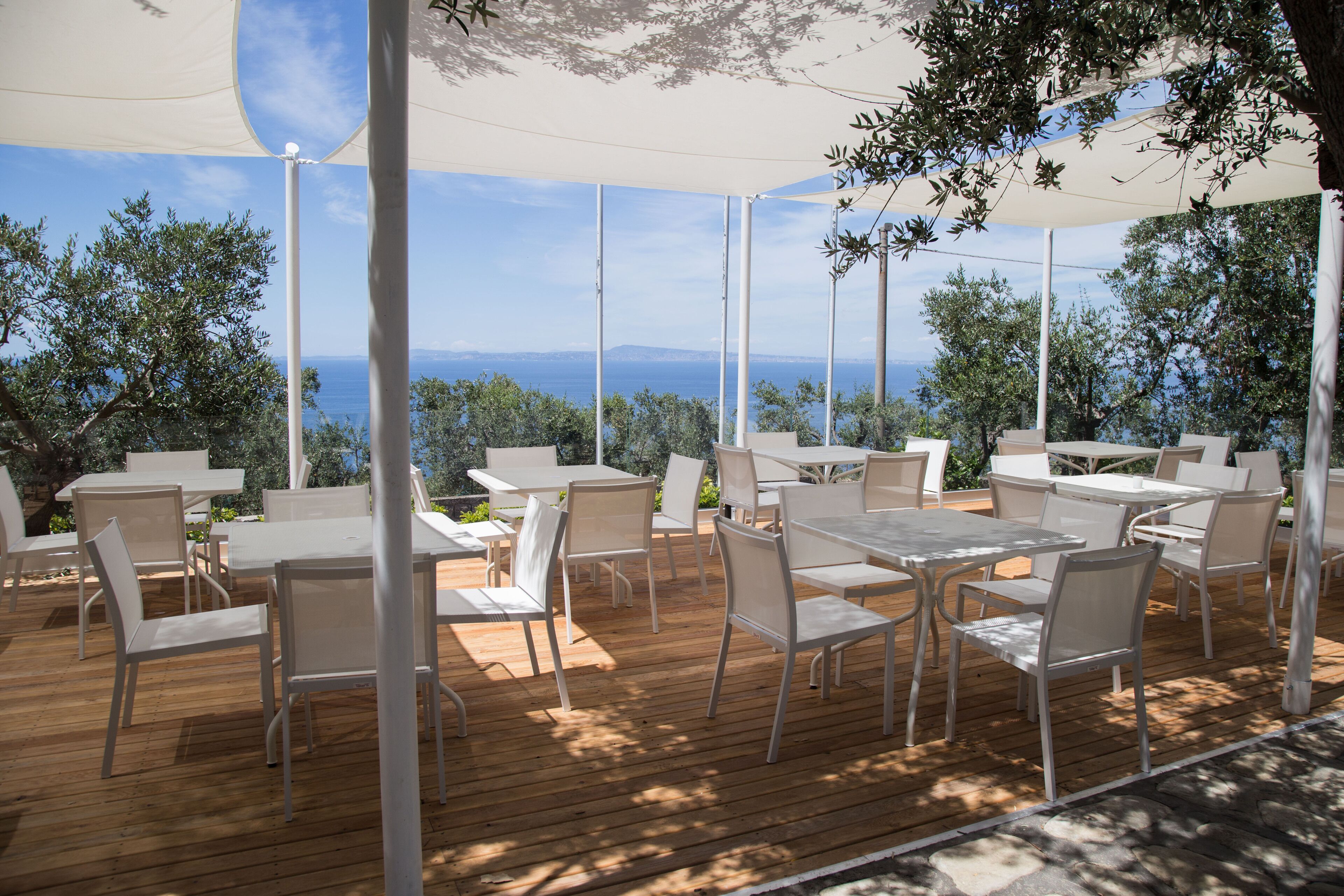 An outdoor seating area with white tables and chairs, overlooking a scenic view of the sea and hills.