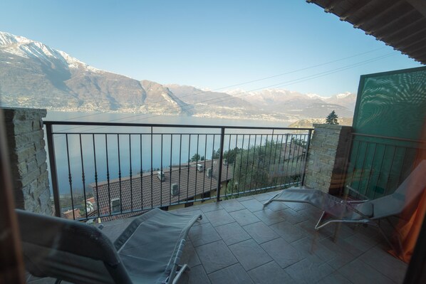 A balcony with a view of a lake and mountains. There are two chairs and a table on the balcony.