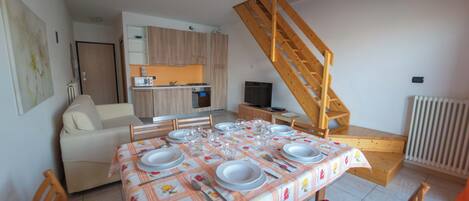 Un comedor con una mesa preparada para cuatro, un sofá, una escalera de madera y una cocina al fondo.