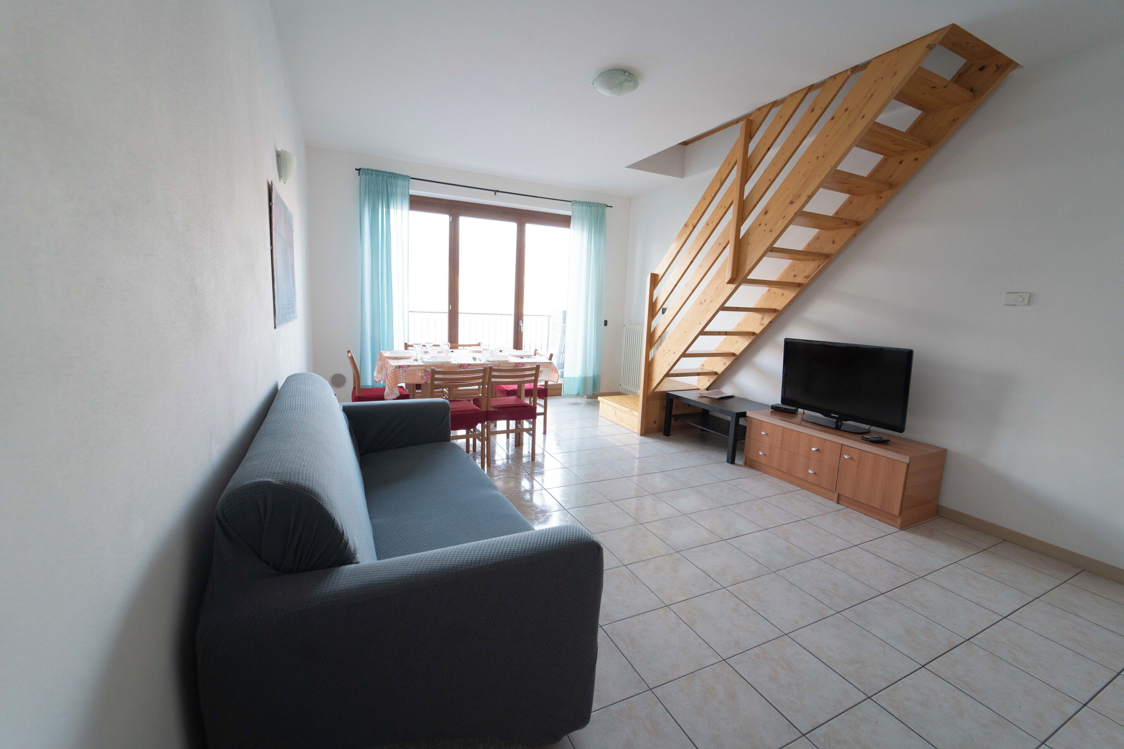 Ein Wohnzimmer mit einer Couch, einem Fernseher auf einem Holzkasten, einem Esstisch mit Stühlen und einer Treppe.