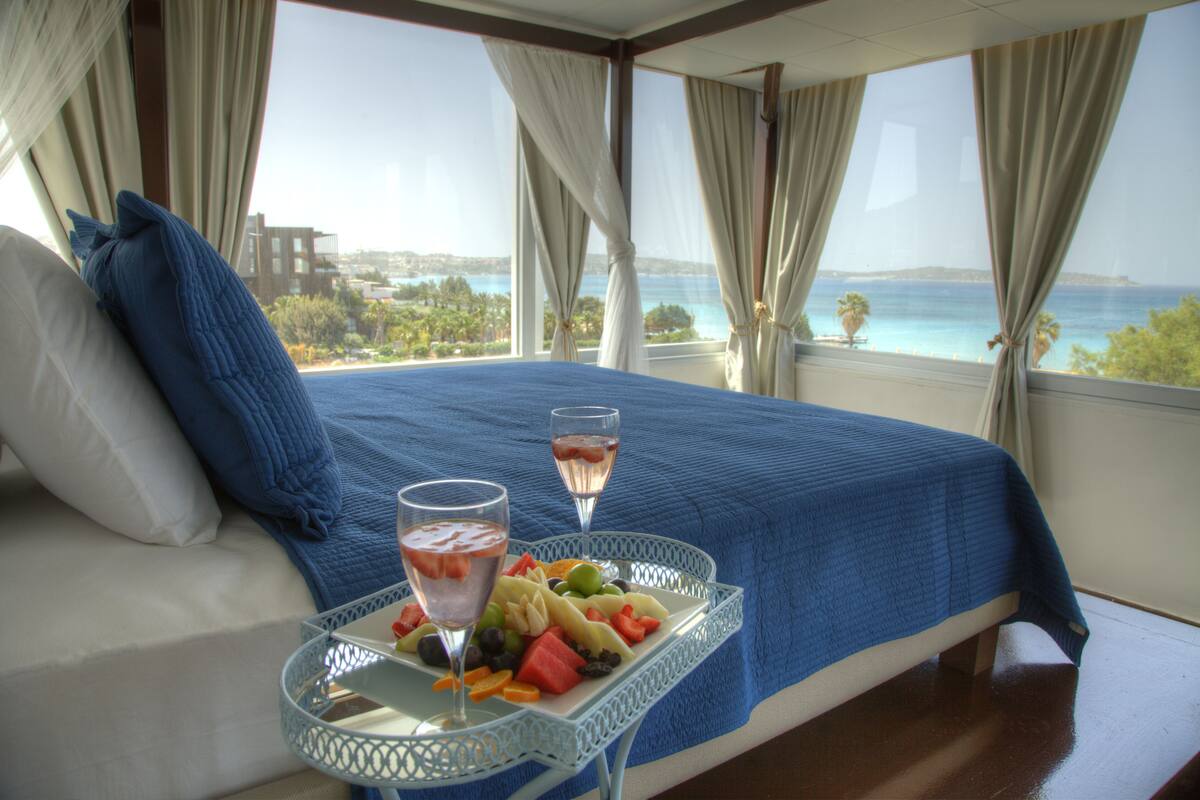 A bed with a blue bedspread, a tray with fruit and two glasses of wine, and a view of the sea through large windows.
