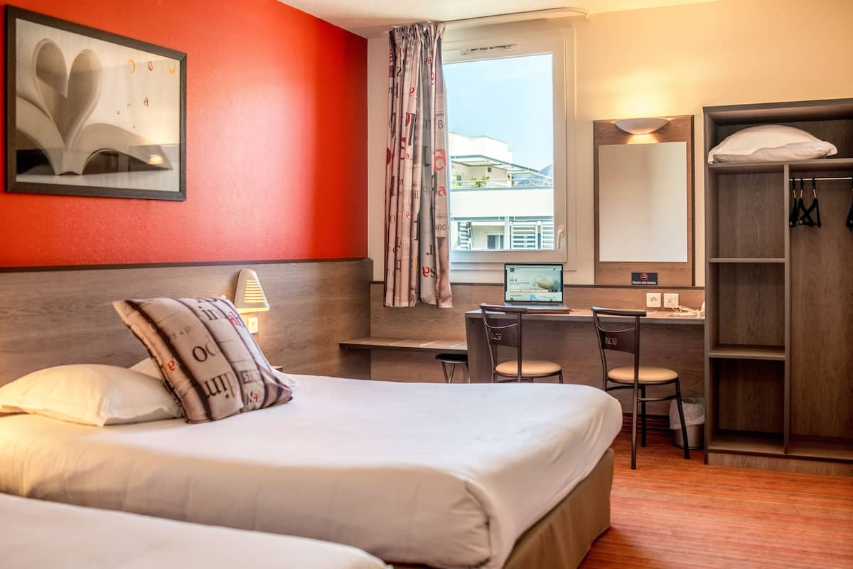 A hotel room with a bed, a desk, a computer, a chair, a wardrobe, and a window with a view of buildings.