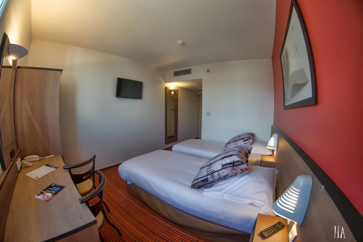 A hotel room with two beds, a desk, a chair, a lamp, and a TV on the wall.