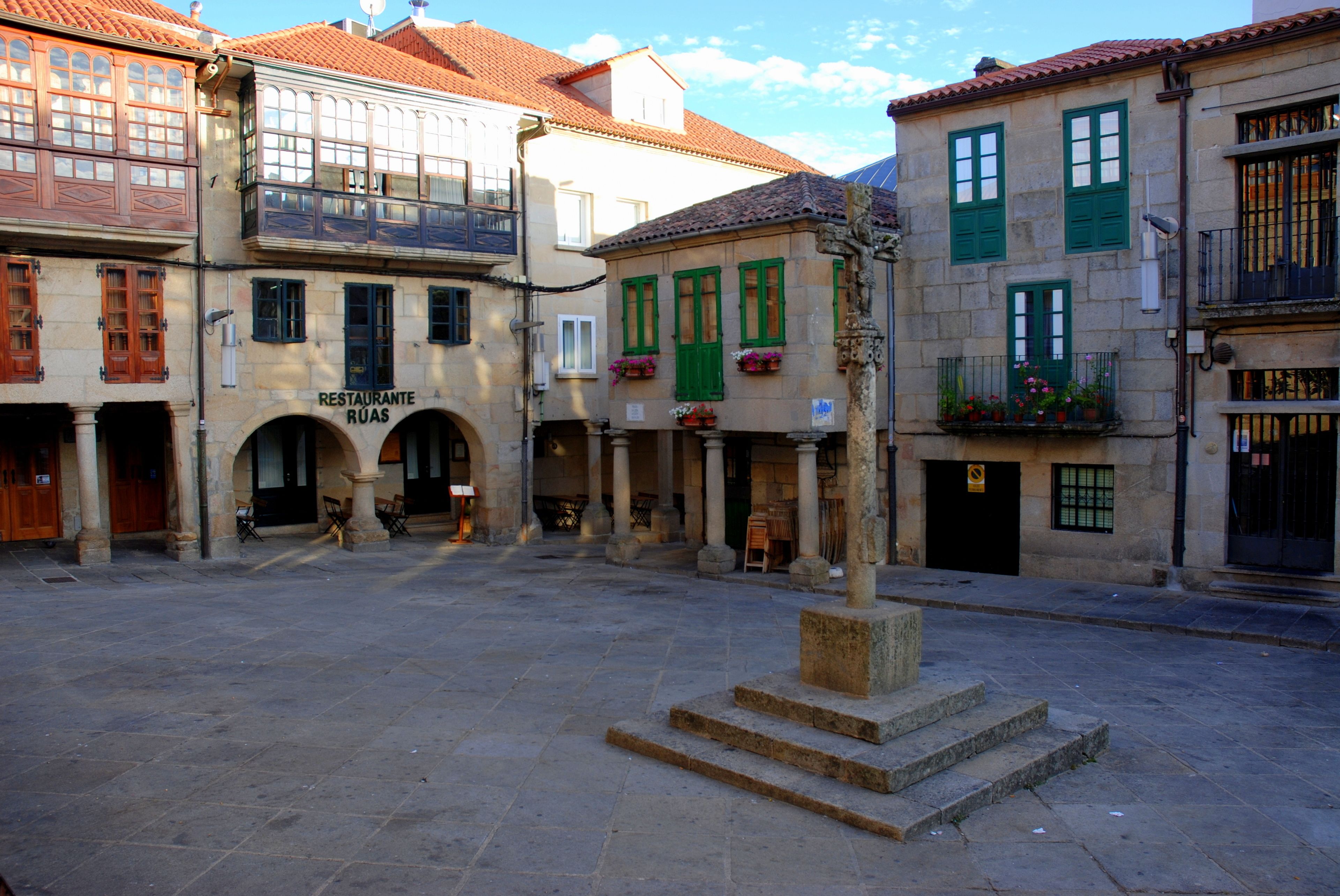 Un patio con suelo de piedra, un monumento central y edificios con puertas arqueadas y contraventanas verdes.