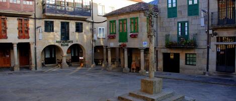 Un patio con suelo de piedra, un monumento central y edificios con puertas arqueadas y contraventanas verdes.