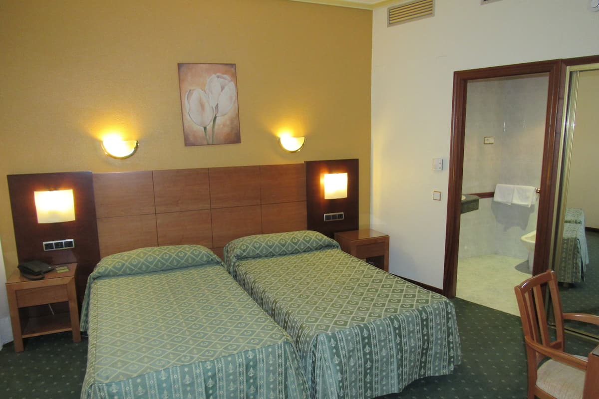 Ein Hotelzimmer mit zwei Betten, einem Nachttisch mit Lampe, einem Gemälde an der Wand und einem durchgegangenen Tür erkennbaren Badezimmer.