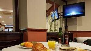 Una mesa de desayuno con cruasanes, fruta, jugo y café, una pantalla de televisión mostrando una escena espacial y una botella de aceite de oliva.