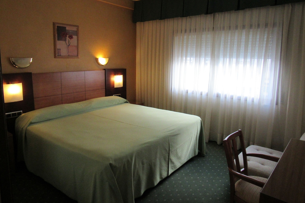 Habitación de hotel con una cama grande, dos lámparas de noche, un cuadro en la pared y una ventana con cortinas traslúcidas.