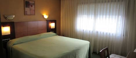 Habitación de hotel con una cama grande, dos lámparas de noche, un cuadro en la pared y una ventana con cortinas traslúcidas.