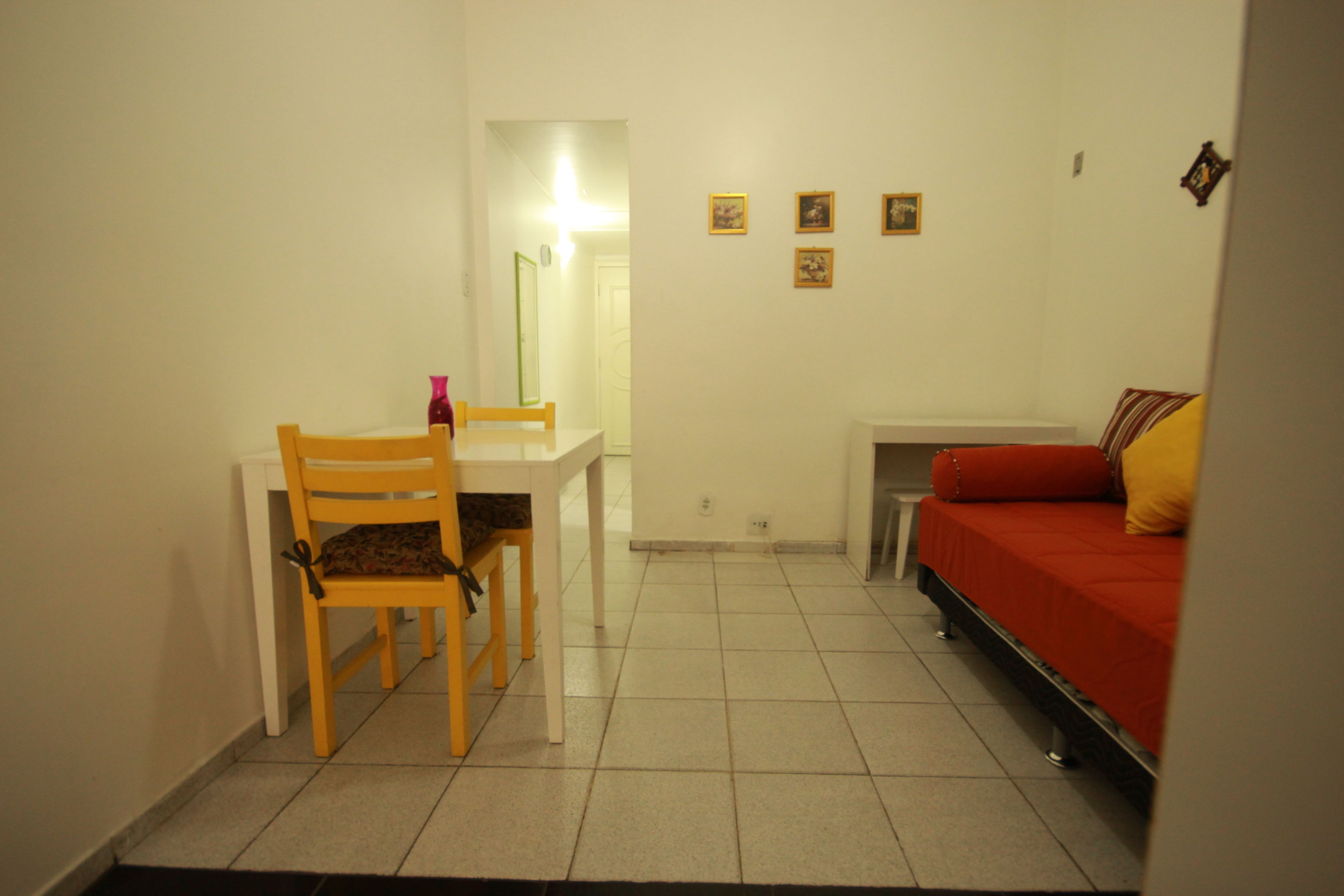 A small, well-lit room with a dining table and chairs, a bed with red bedding, and a few framed pictures on the wall.