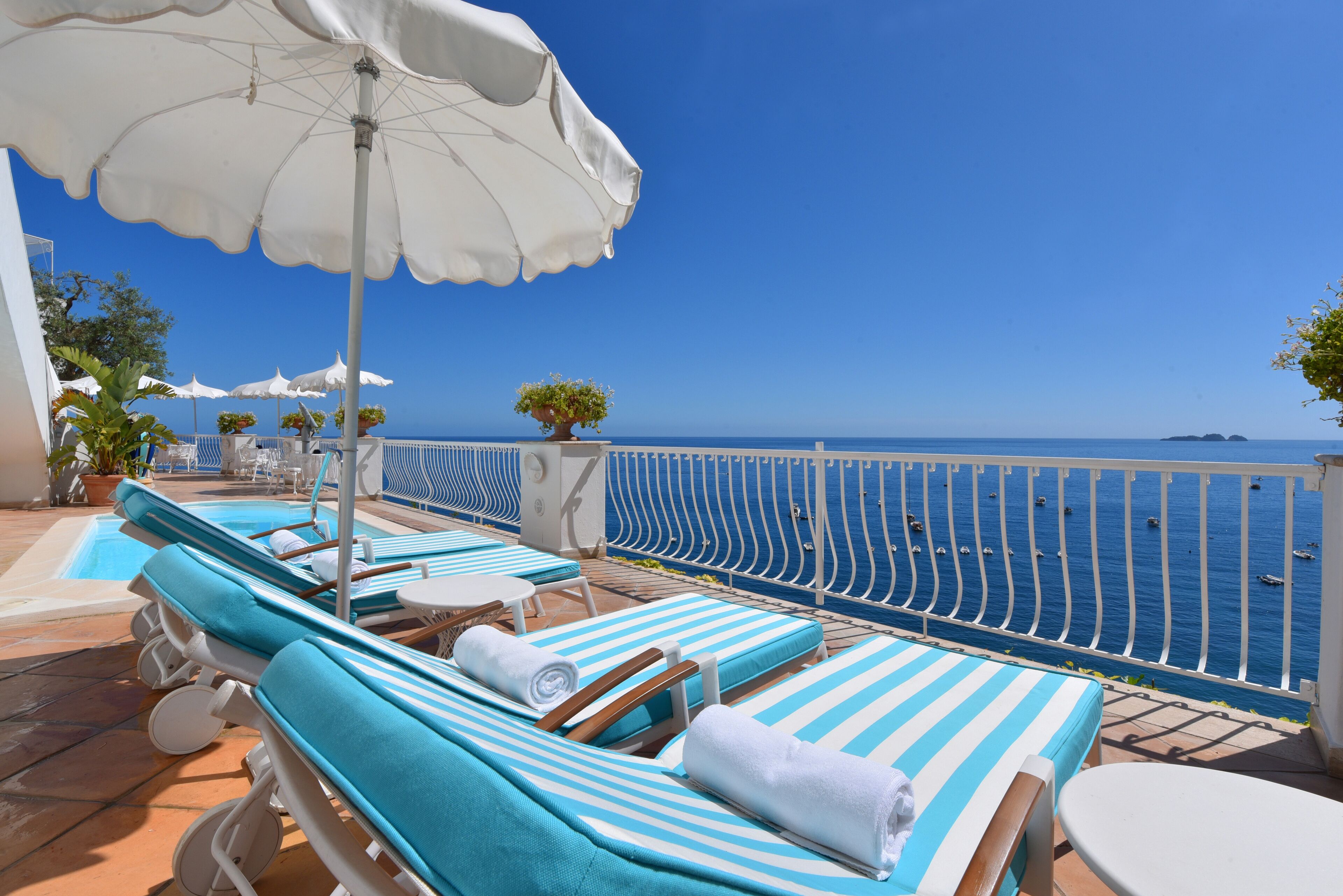 A sunlit outdoor lounge area with striped blue and white lounge chairs, white umbrellas, and a view of the sea.