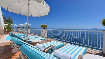 A sunlit outdoor lounge area with striped blue and white lounge chairs, white umbrellas, and a view of the sea.