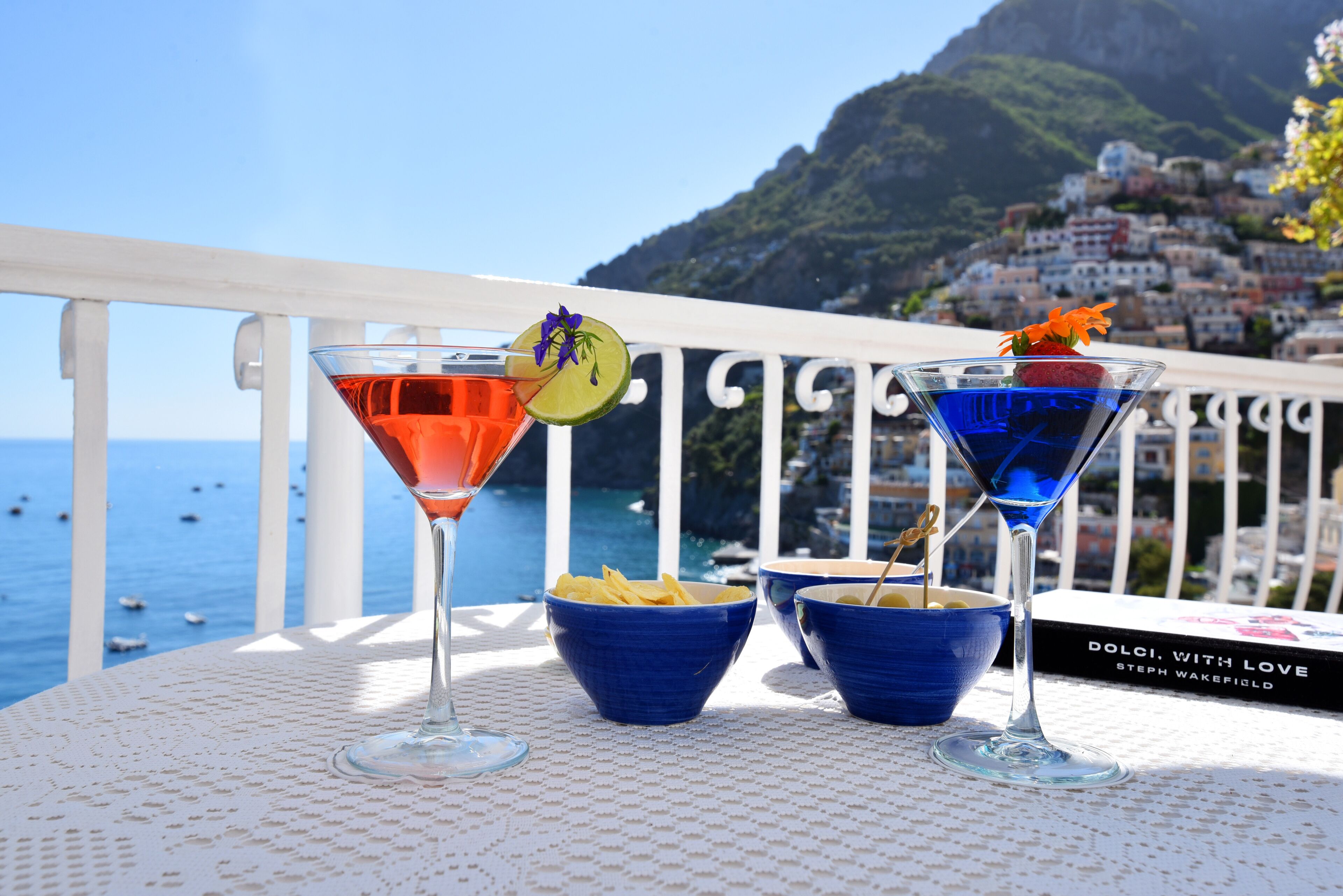 Une table avec deux verres à martini, un livre intitulé « Dolci, avec amour », et des bols de chips, le tout sur un balcon offrant une vue sur la mer et une ville perchée sur une colline.