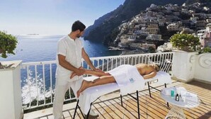 Iemand ontvangt een massage op een balkon met uitzicht op een kustplaatsje onder een heldere lucht.