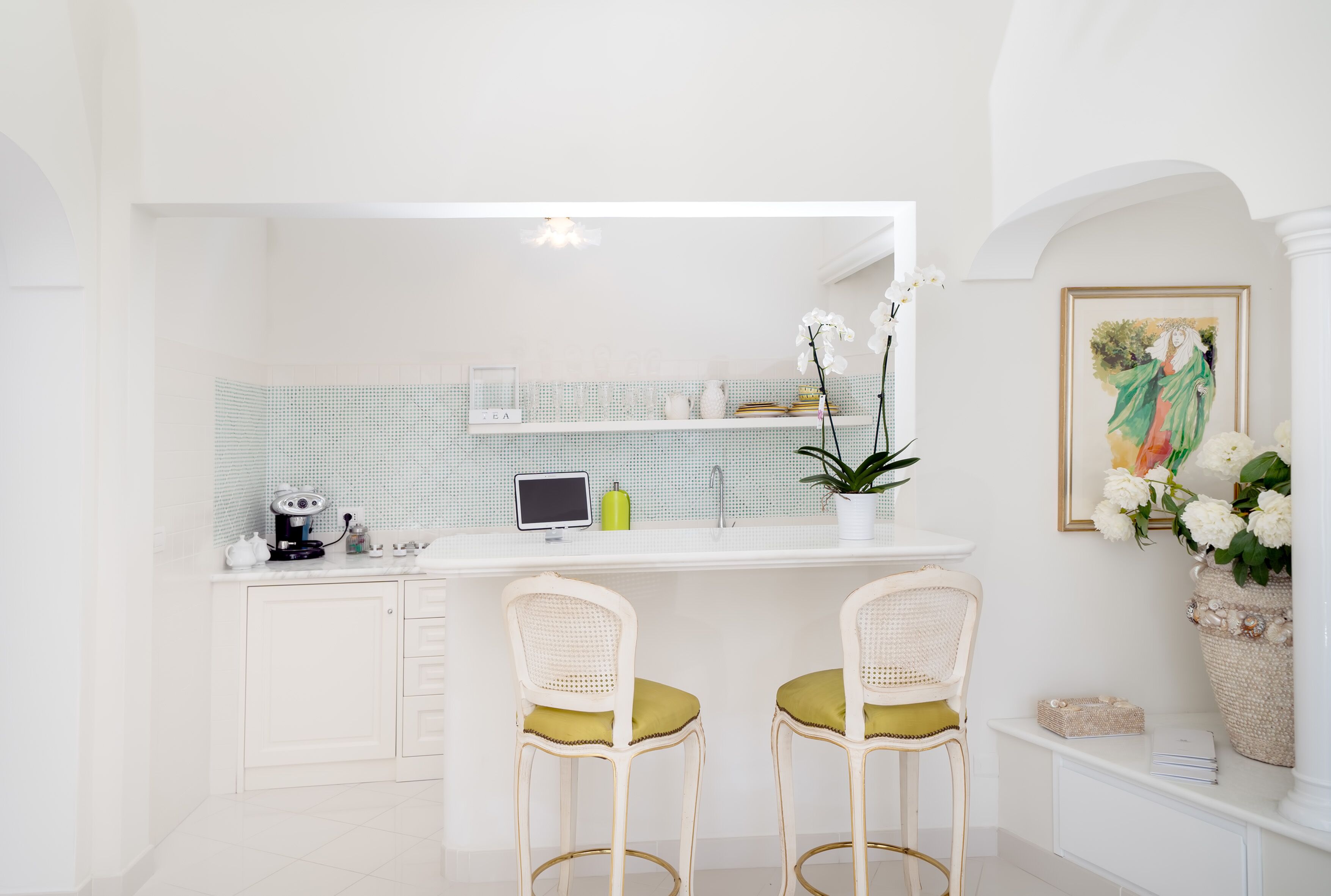 A modern kitchen with white cabinetry, a white countertop, two white wicker chairs with green cushions, a small island with a tablet, a coffee machine, and a potted plant.
