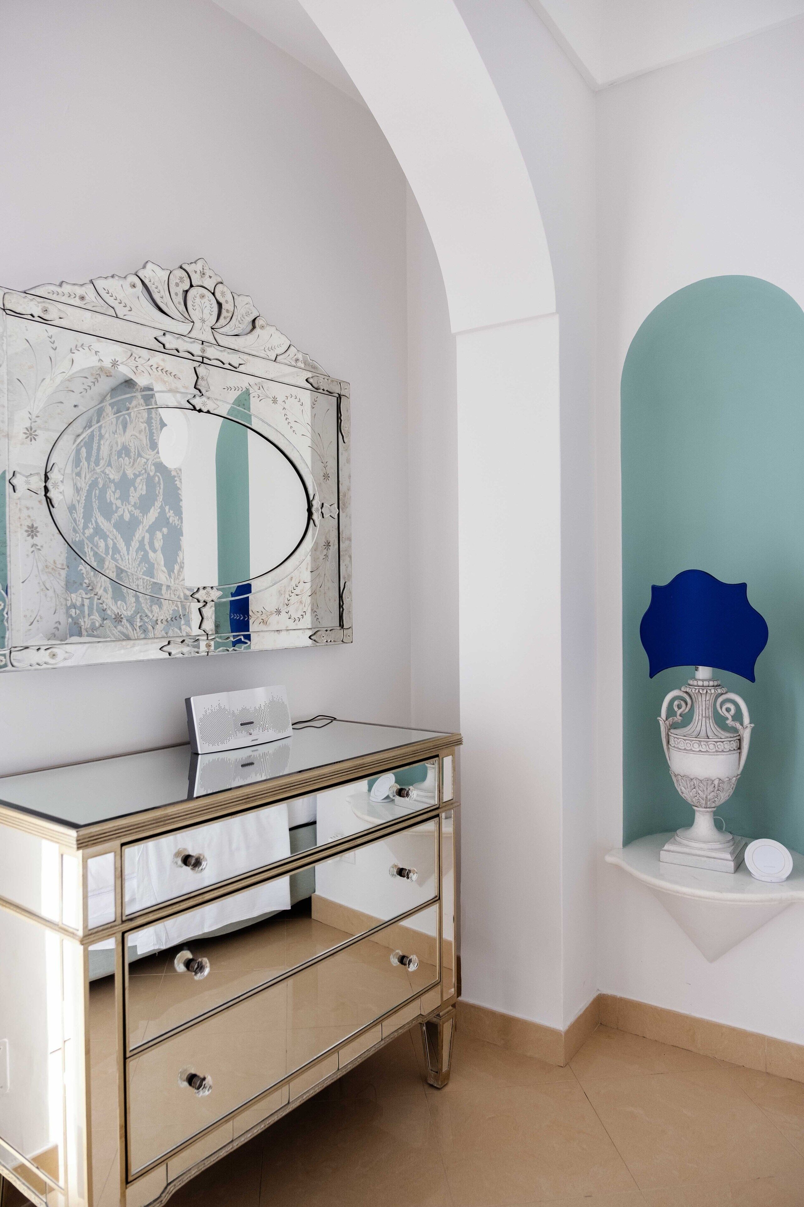 Ein Zimmer mit einer Spiegelwand, einem weißen Schminktisch mit Schubladen, einem kleinen Regal mit Dekoration und einer blauen Akzentwand.