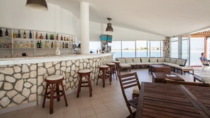 Et rummeligt område indendørs med en bar i natursten, flere træbjerke, et stort opholdsområde med sofaer, og havudsigt gennem store vinduer.