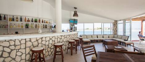 A spacious indoor area with a stone wall bar, multiple wooden stools, a large seating area with sofas, and a view of the sea through large windows.