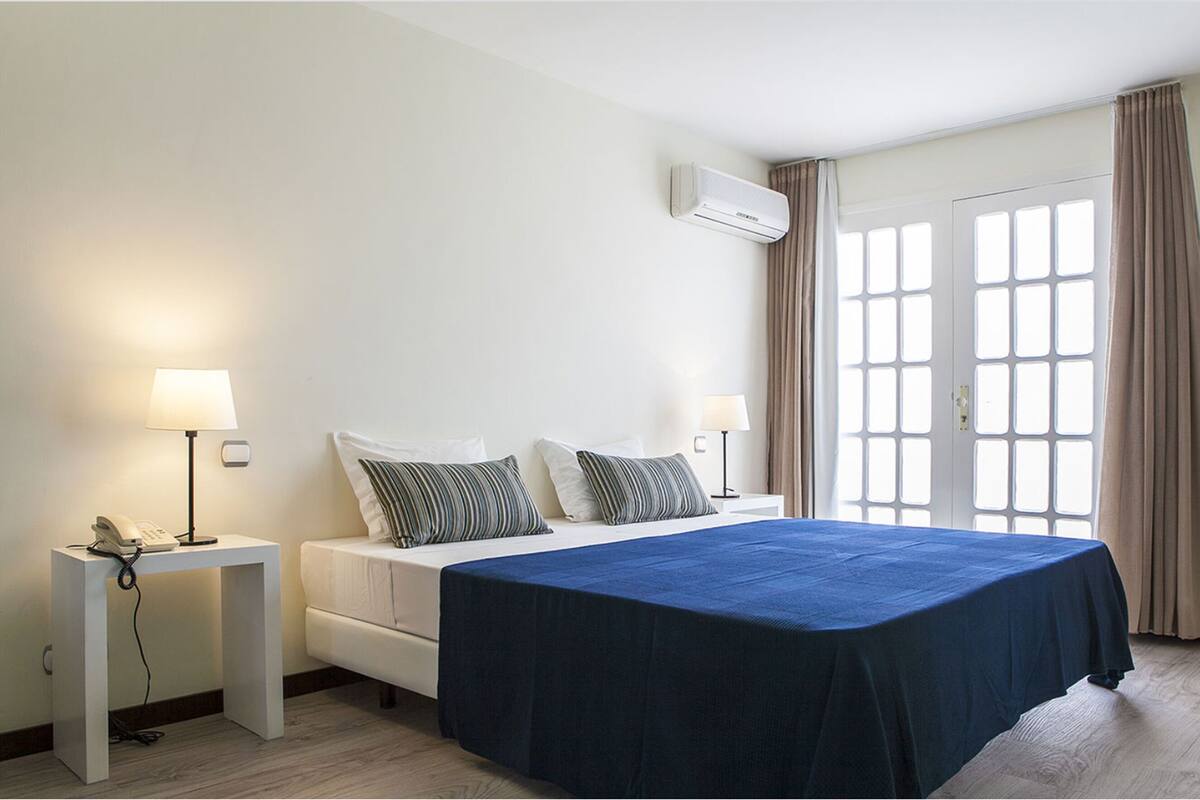 Ein Hotelzimmer mit Bett, Nachttischen, Lampen, einer Klimaanlage und einem Fenster mit Vorhängen.