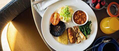 Un plato blanco con una variedad de alimentos, incluyendo un tomate a la parrilla, frijoles, verduras de hoja verde y un trozo de pollo a la plancha, acompañado de un vaso de jugo de naranja y un tarro de bayas.