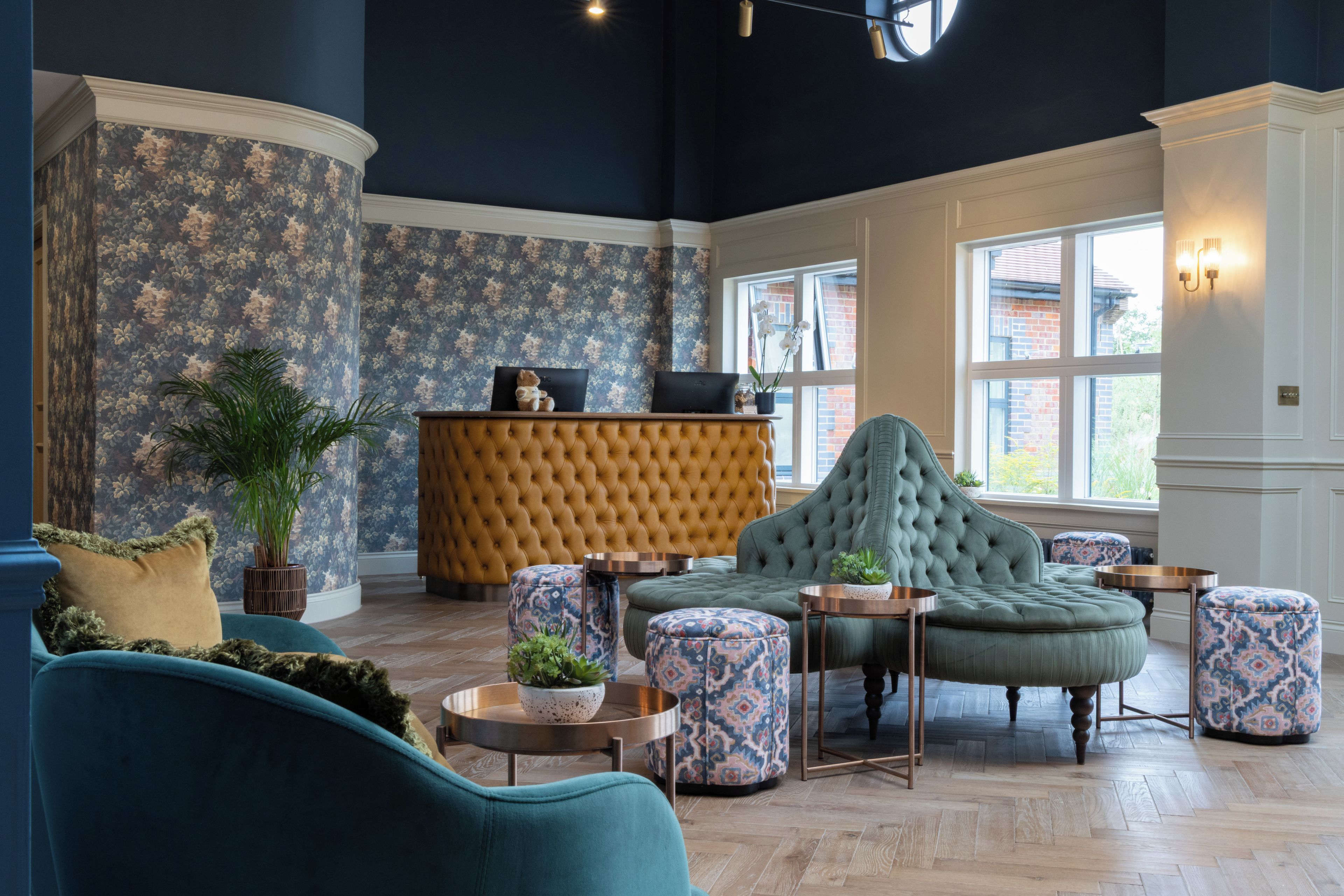 Eine Lobby mit einer gepolsterten Couch, gemusterten Hocker, einem Empfangstresen und einem großen Fenster.