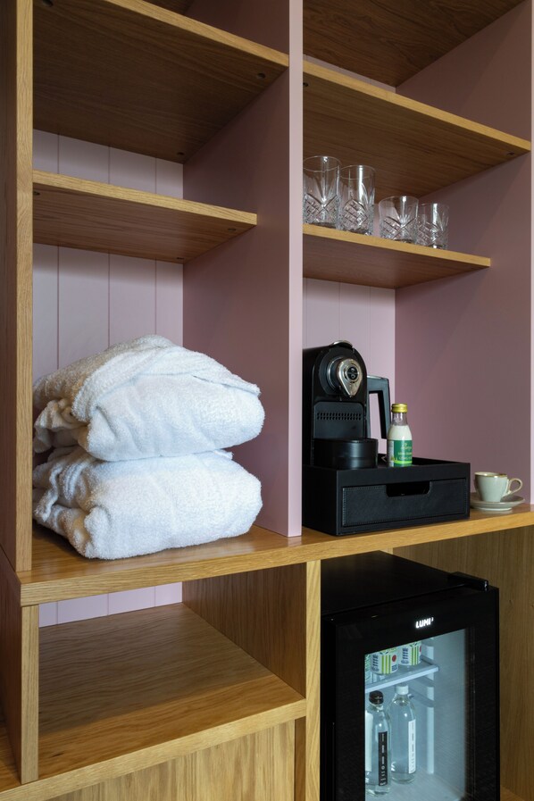 A hotel room with wooden shelves, a stack of white towels, a coffee machine, a glass of milk, and a refrigerator. - Bernwood Park Hotel (Aylesbury)