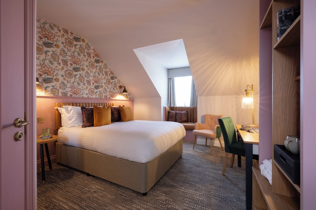 A hotel room with a bed, a desk, a chair, a lamp, and a window.