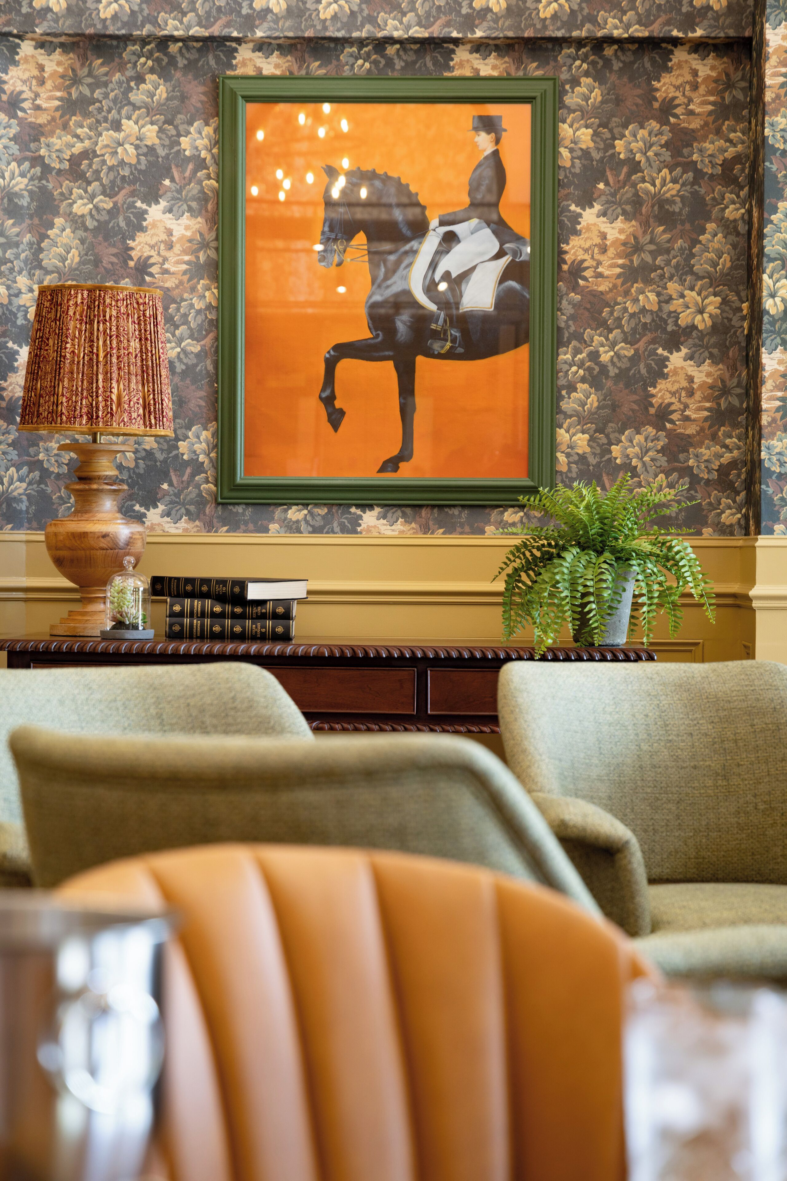 A room with a framed artwork of a horse, a lamp, books, and a potted plant.