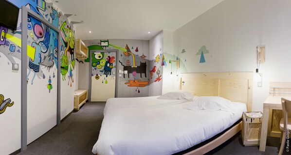 A hotel room with a large bed, a wooden headboard, a bedside table, and a nightstand. There is a door leading to another room and a wall with colorful artwork. - Hotel Graffalgar (Strasbourg)