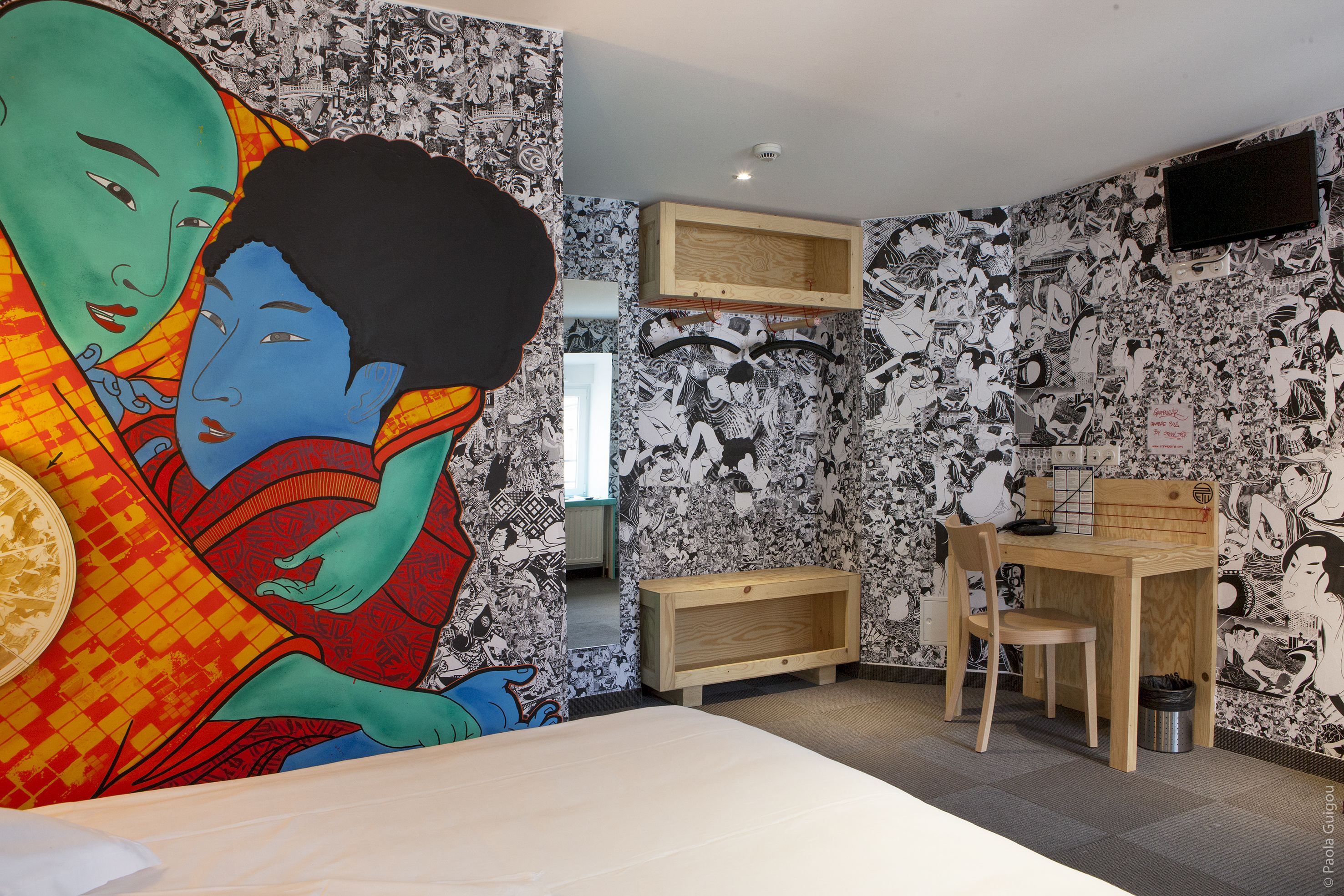 Une pièce comprenant un lit, un bureau et une chaise. Un des murs est recouvert d’un papier peint à motifs noir et blanc.