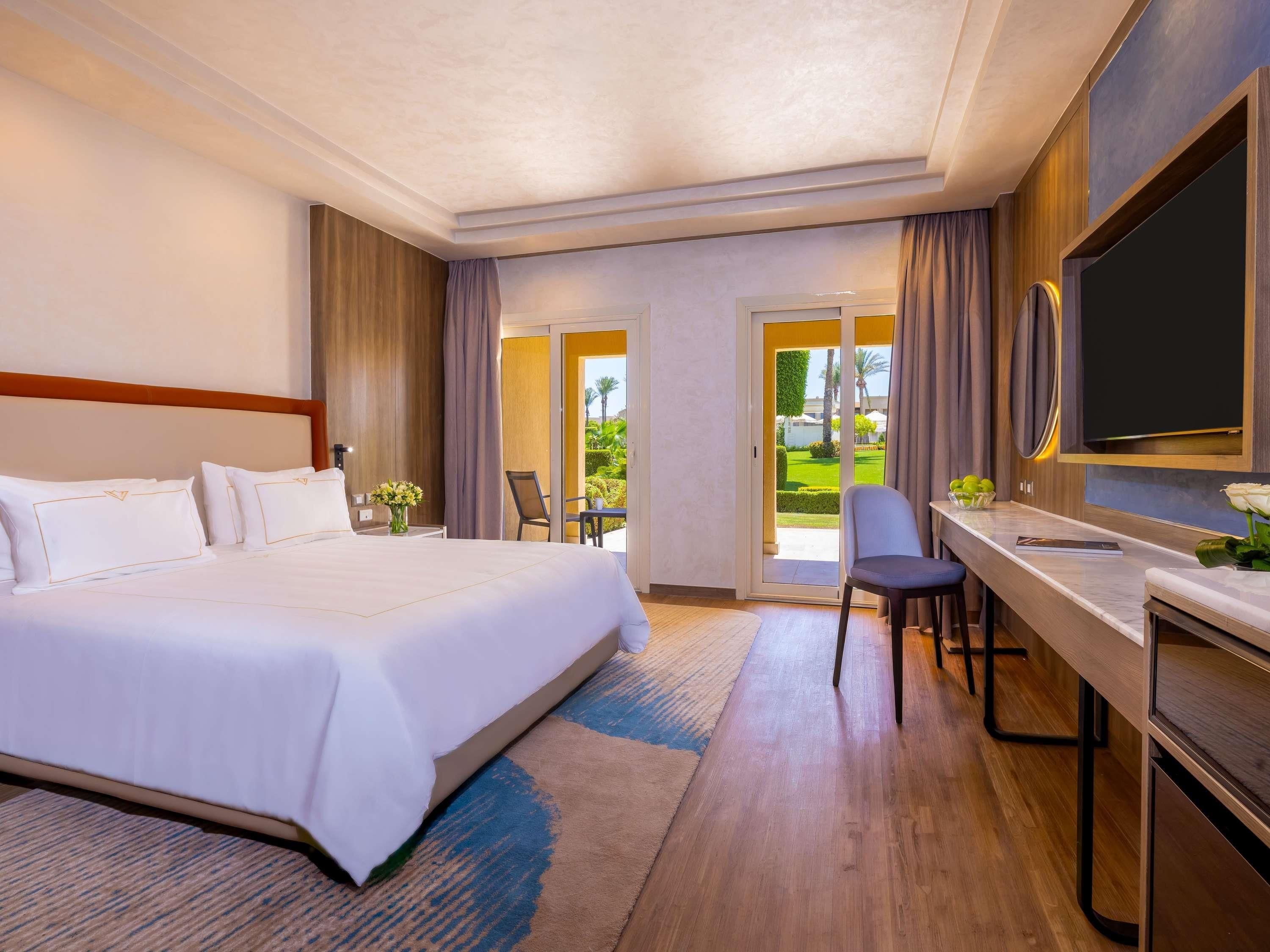 Une chambre d’hôtel avec un grand lit, un bureau avec une chaise, une télévision et une vue sur un jardin depuis la porte.