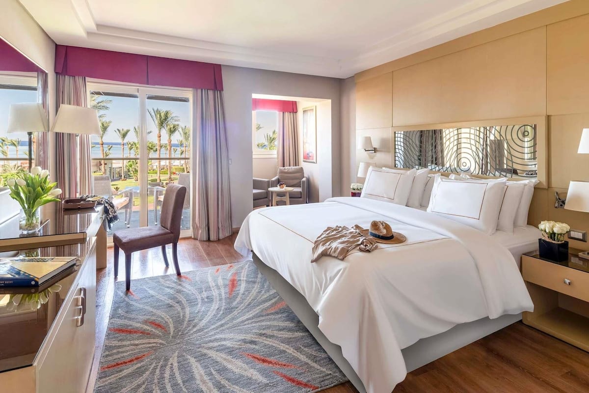 A hotel room with a bed, a desk, a chair, a view of the ocean, and a balcony.