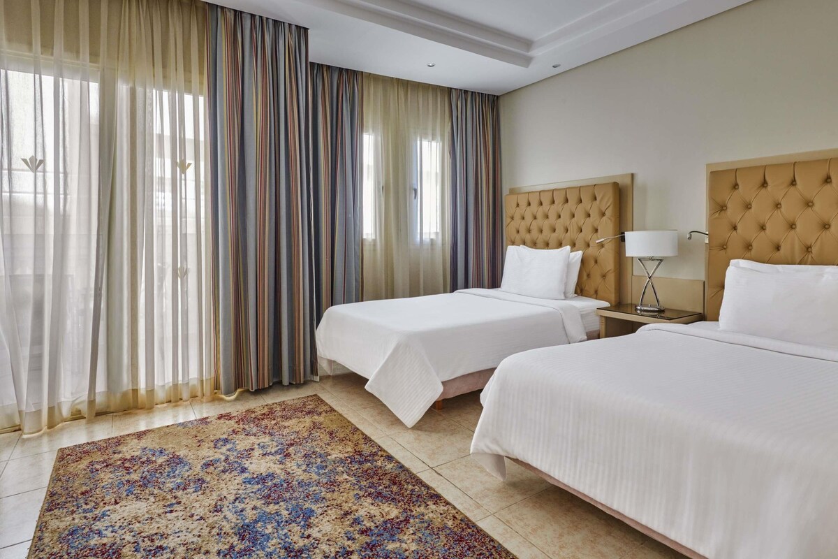 A hotel room with two beds, a large window with sheer curtains, a patterned rug, and a nightstand with a lamp.