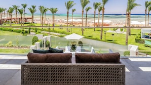 A balcony with wicker furniture, a glass table, and a view of a beach with palm trees and umbrellas. - Rixos Premium Seagate (Sharm El Sheikh)