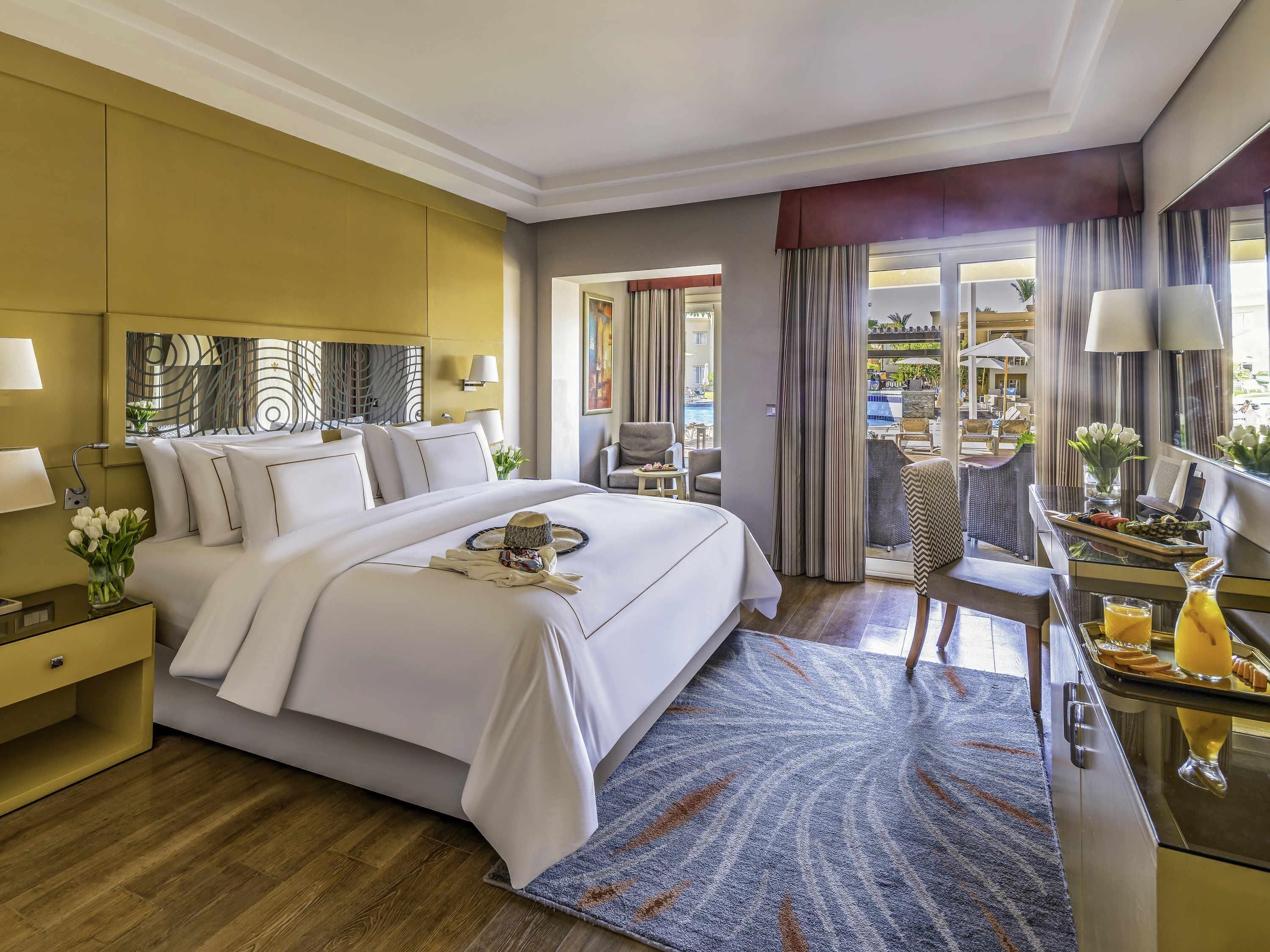 A hotel room with a large bed, a desk with a tray of breakfast, a chair, and a view of the outside.