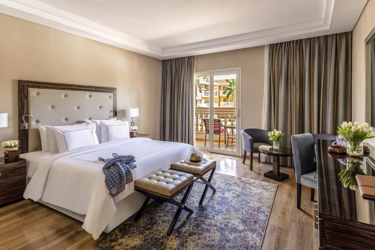 A hotel room with a large bed, a desk, a chair, a small table, and a view of the outside.