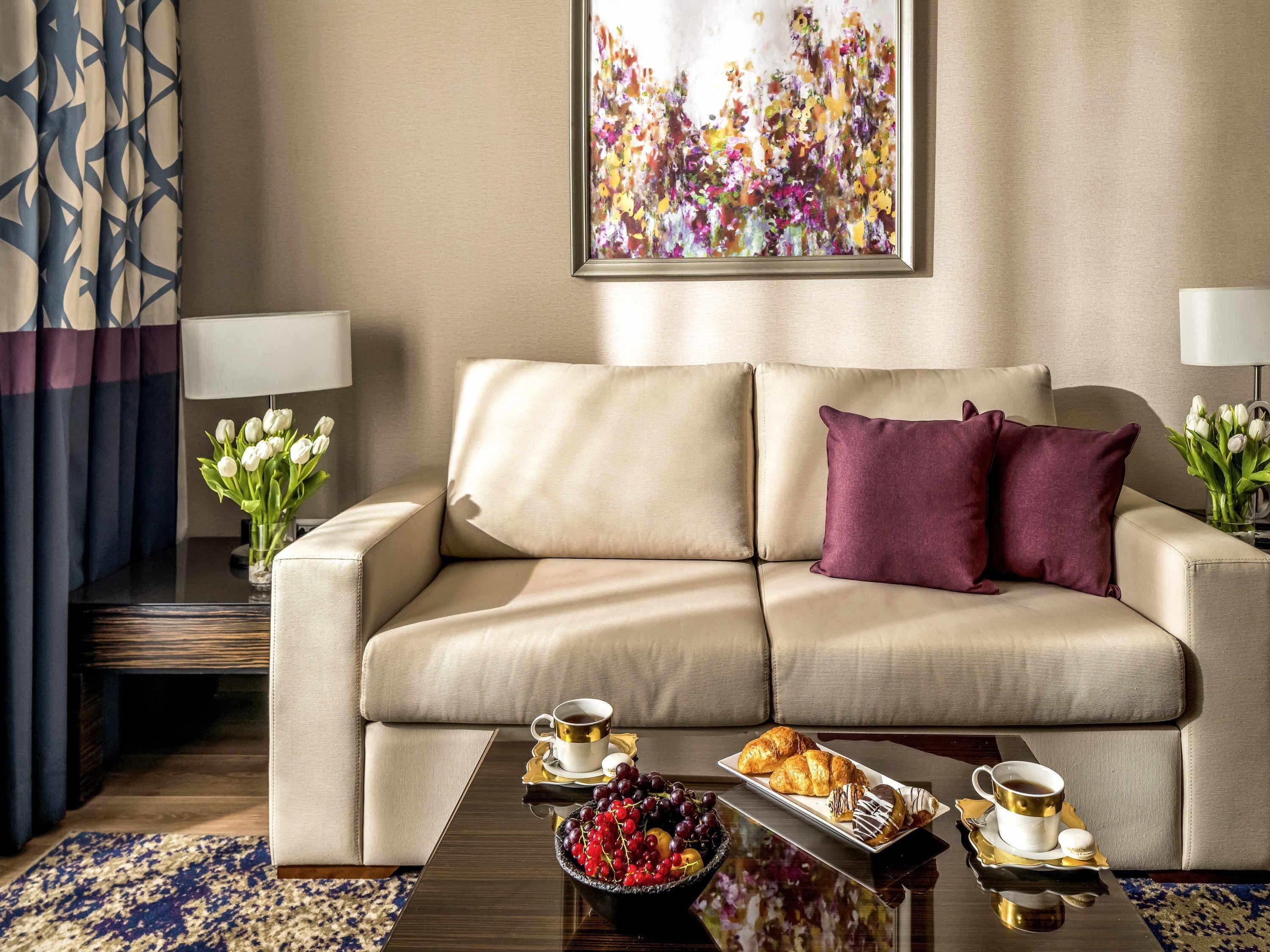 Een moderne woonkamer met een beige bank, een bijzettafeltje met gebakjes en een fruitschaal, en een schilderij aan de muur.