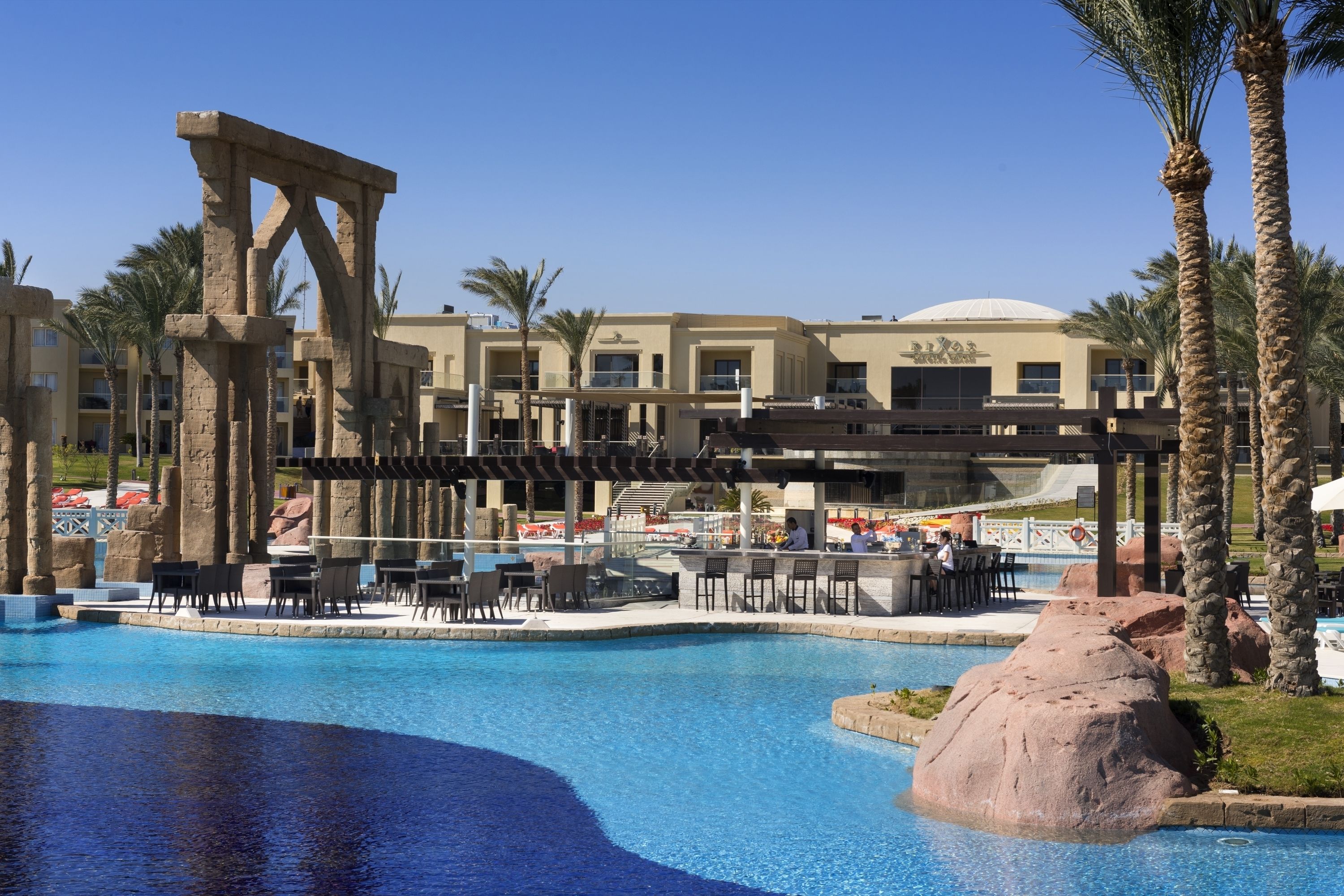 A pool area with a large rock centerpiece, palm trees, and a building with a dome in the background.