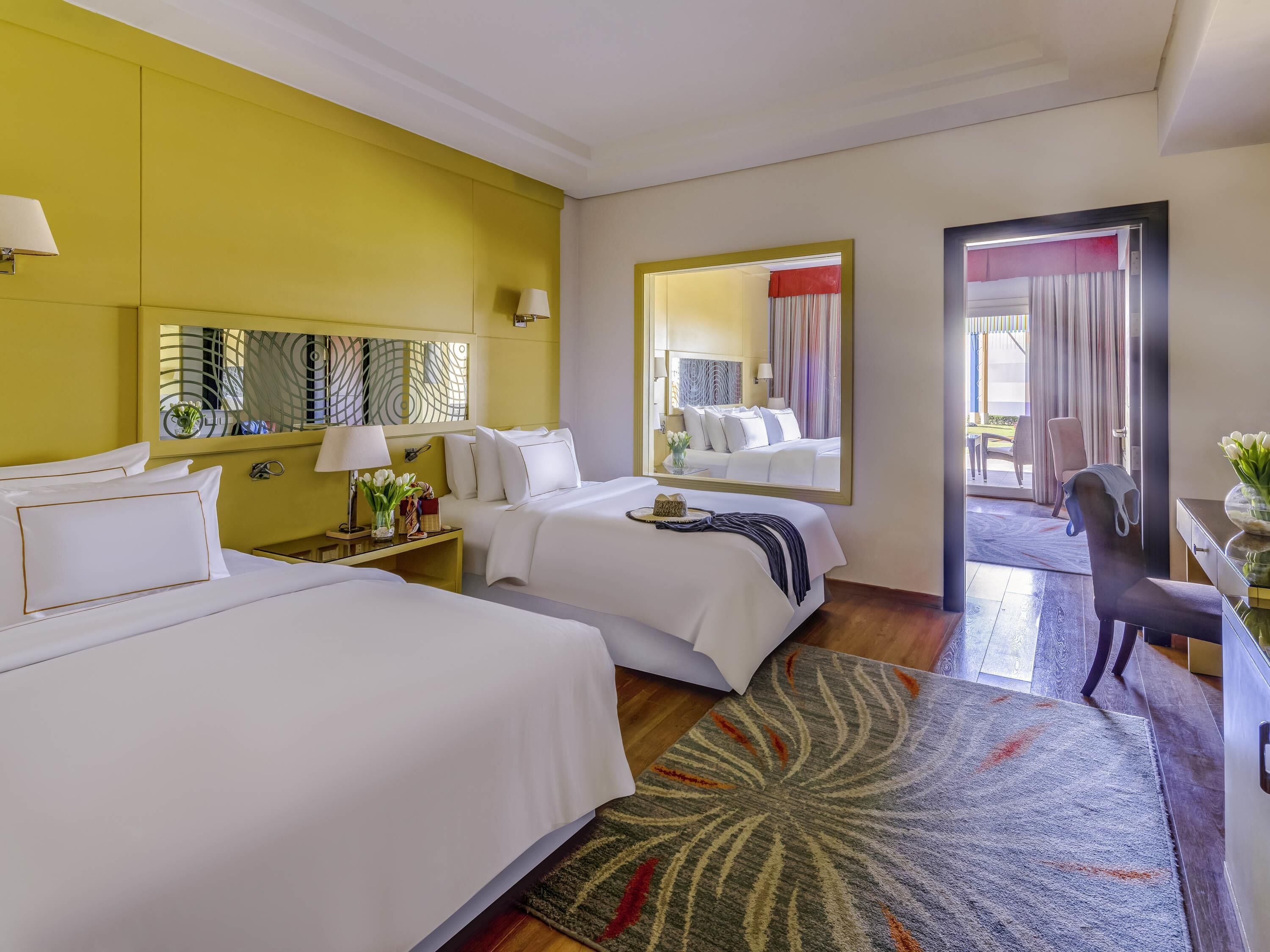A hotel room with two beds, a large mirror, a nightstand with a lamp, and a view of the outside.