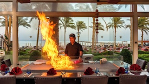 A chef performing a fire act at a table with a view of a beach and palm trees. - Rixos Premium Seagate (Sharm El Sheikh)