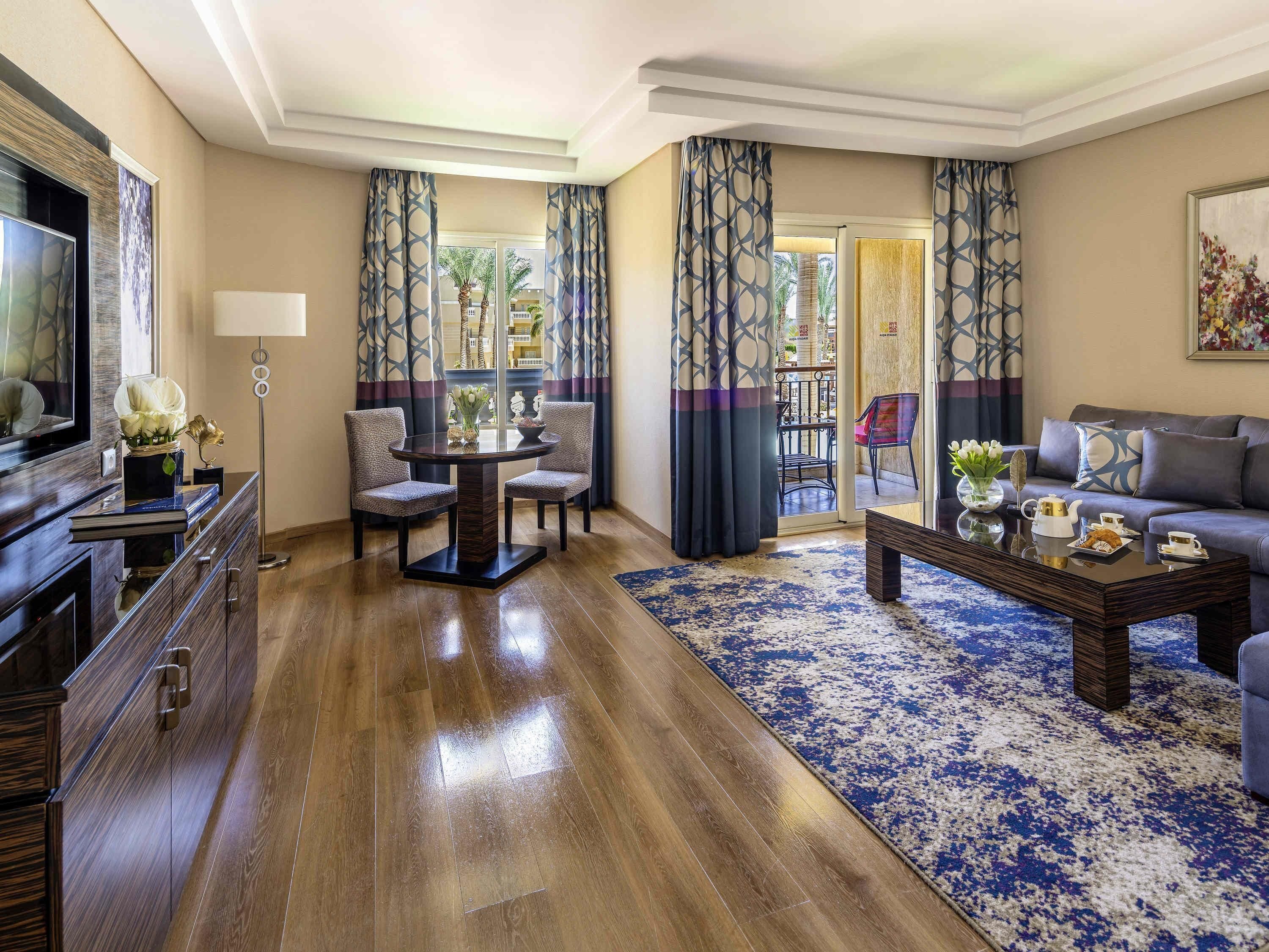 A modern living room with a wooden floor, a round table with chairs, a sofa, a coffee table, and a TV on a stand.