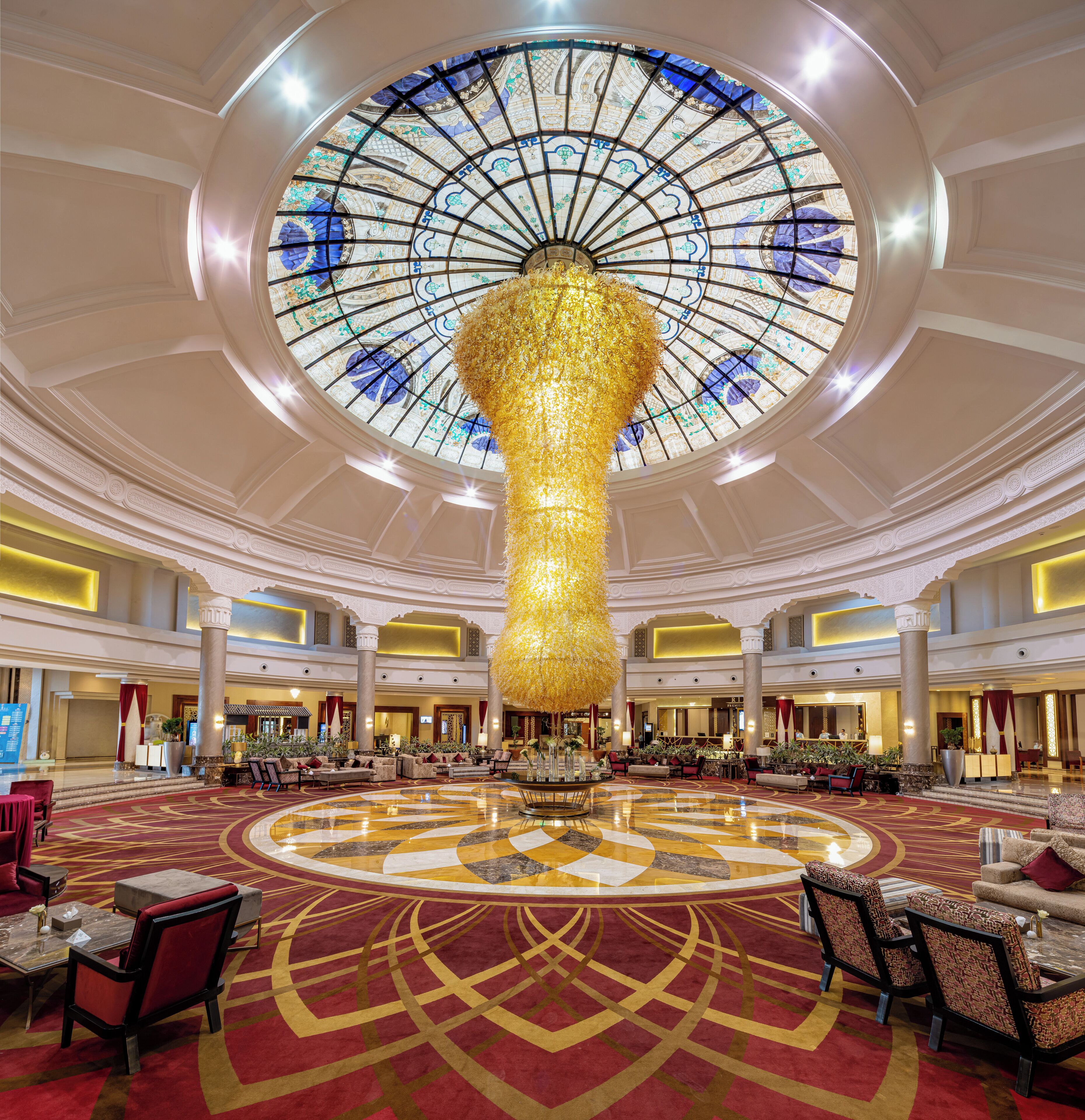 A grand lobby with a large, ornate chandelier, a circular patterned carpet, and a high ceiling with a glass dome.