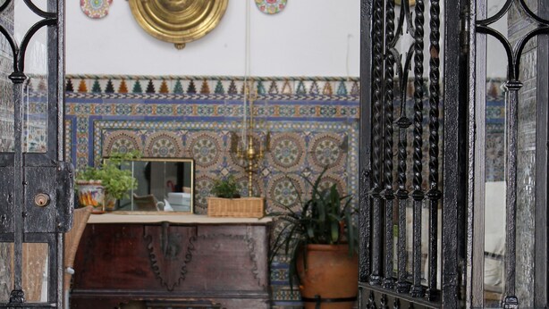 An open wrought iron gate leading to a room with a tiled wall, a small table with a potted plant, and a decorative plate on the wall. - Pensión El Portillo (Córdoba)