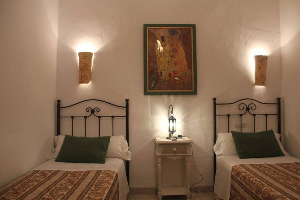 A room with two beds, each with a green pillow and a patterned bedspread. There is a painting on the wall and two wall-mounted lamps.