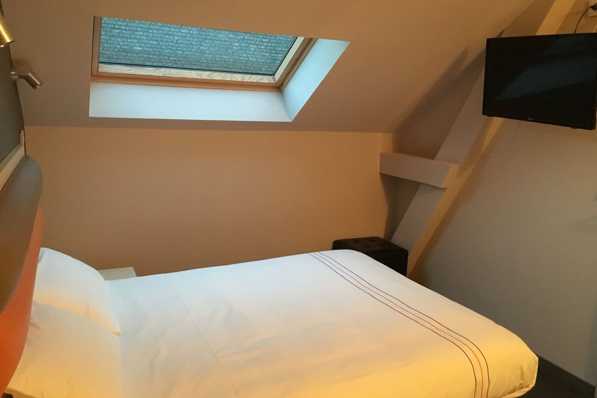 A compact room with a skylight, a bed, a small bedside table, and a mounted TV.