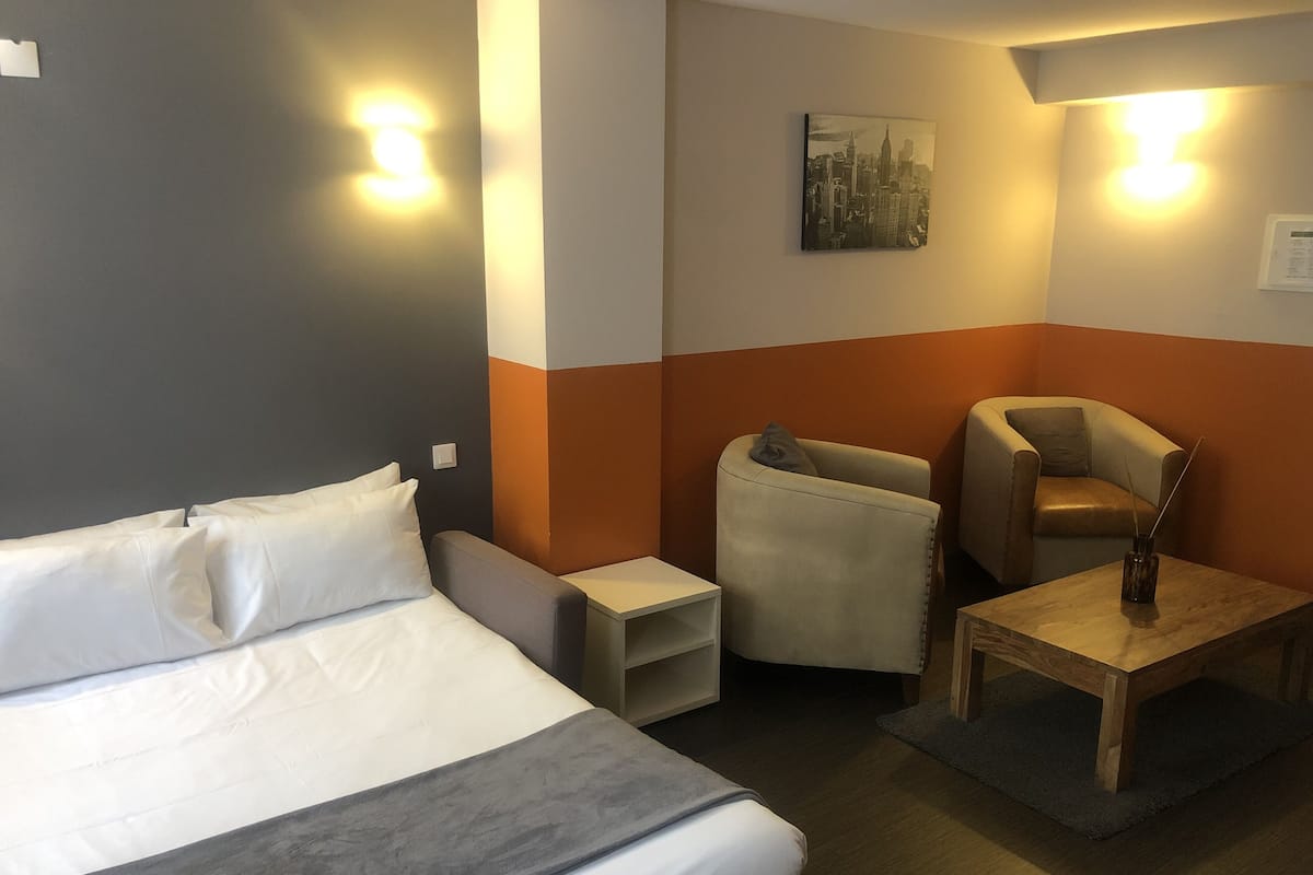 A hotel room with a bed, a nightstand, a chair, a small table, and a wall-mounted picture.