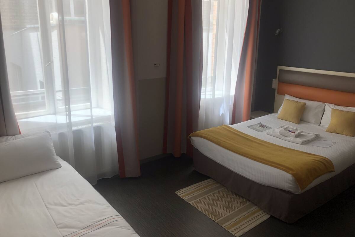 A hotel room with a bed, a bedside table, a window with sheer curtains, and a view of the outside.