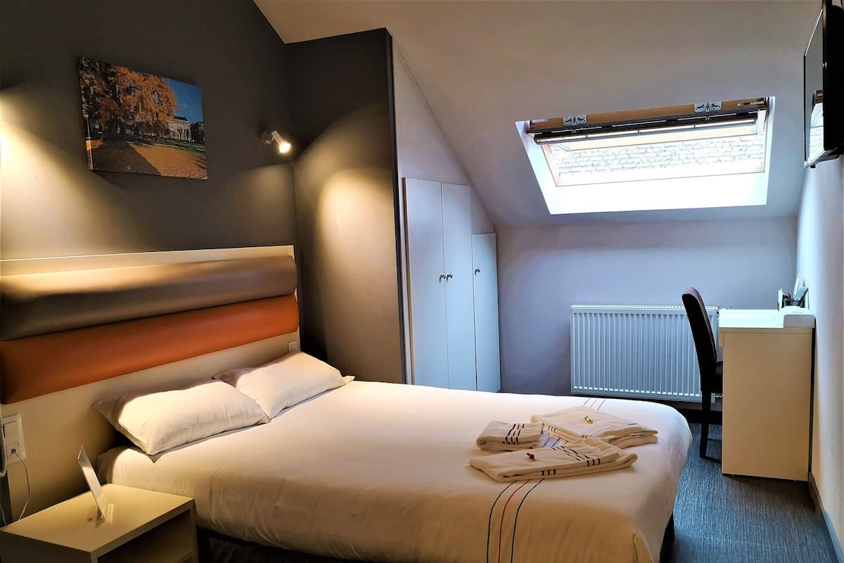 A modern hotel room with a neatly made bed, a desk with a chair, a radiator, and a skylight.