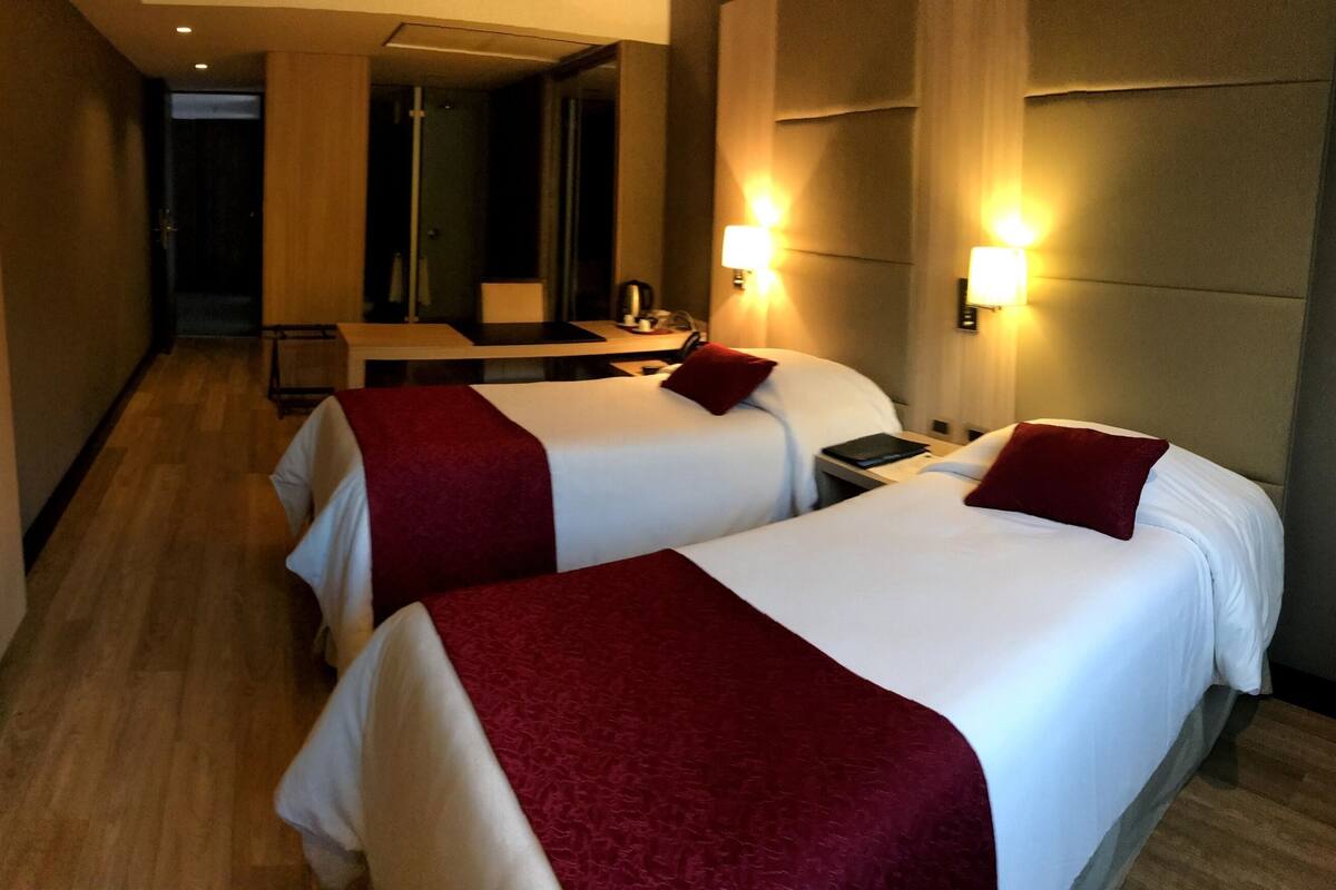 A hotel room with two beds, a mirror, a desk, and a bathroom visible in the background.