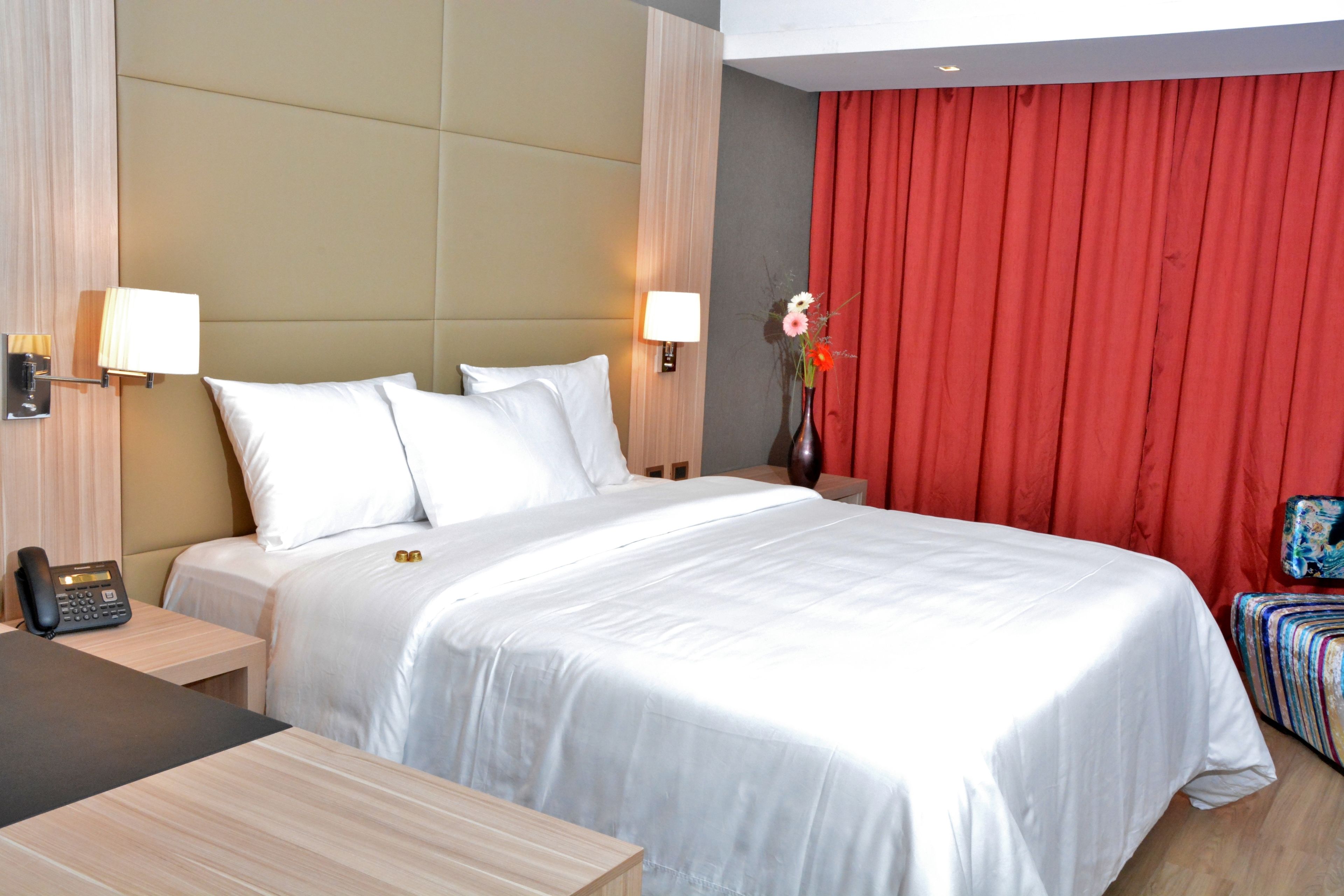 A hotel room with a large bed, two bedside tables, a desk, a chair, and a red curtain.